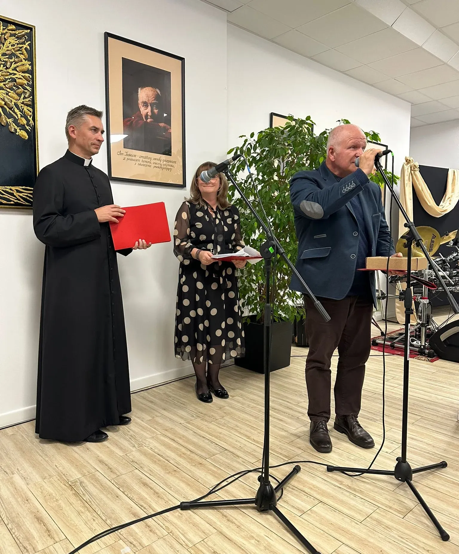 Jasne pomieszczenie. Trzy osoby stoją na jasnej podłodze. Mężczyzna po prawej mówi do mikrofonu, trzymając opakowanie w rękach. Po lewej stronie stoi ksiądz w czarnej sutannie z czerwoną teczką. Między nimi stoi kobieta w czarnej sukni w beżowe grochy, trzyma dokumenty. Za nimi wysoka roślina doniczkowa, na ścianie wiszą dwa obrazy, w tym portret starszego mężczyzny.